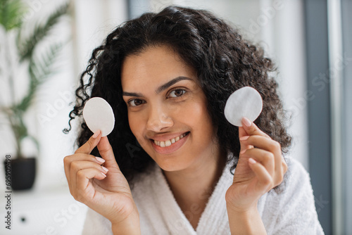 Close-up portrait of positive curly haired multinational woman in bathrobe with clean smooth flawless skin, removing make-up with cotton pads, spa, therapy, treatment, on background of bright bedroom.