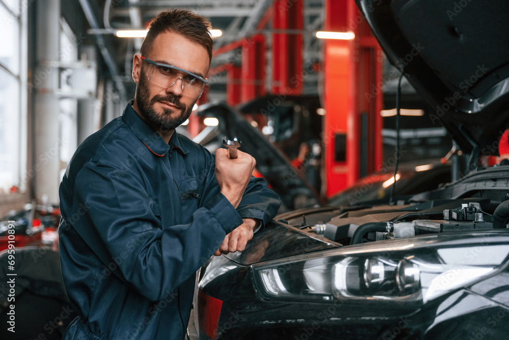 Looking at the camera. Car repairman is in the garage with automobile Looking at the camera. Car repairman is in the garage with automobile