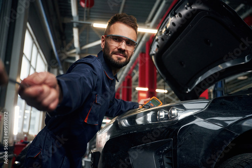 Looking at the camera. Car repairman is in the garage with automobile