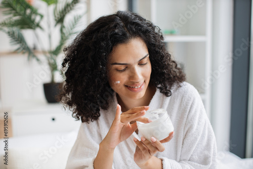 Beautiful African American woman holding jar and applying cream on her face with healthy skin while posing on background of light bedroom. Curly afro hair youth and skin care concept. Beauty face.
