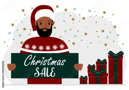 A man in a sweater and a red Santa hat holds a poster with the text about Christmas sale in his hand.