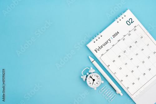 February 2024, Monthly desk calendar for 2024 year and alarm clock with paper clips on blue color background.