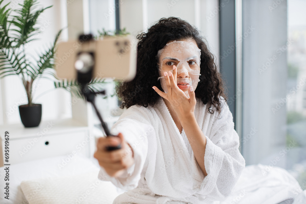 Pretty African female in white bathrobe vlogging using phone on selfie stick in white bathrobe touching face with applied paper sheet mask during cosmetic online video class at home. Pretty African female in white bathrobe vlogging using phone on selfie stick in white bathrobe touching face with applied paper sheet mask during cosmetic online video class at home.