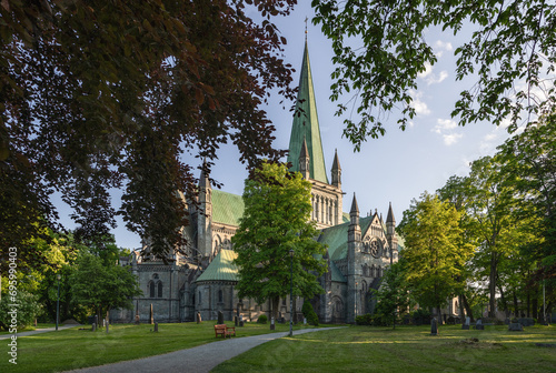 Sunset view of Nidaros Cathedral (Nidarosdomen) framed by lush green leaves in Trondheim, Norway, against a clear blue sky on a summer evening