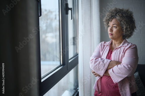 A mature woman of mixed race looking outside a large window, her expression reflecting sadness, concern, and a sense of depression photo
