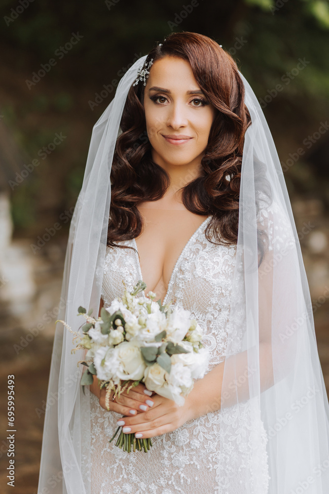 Curly brunette bride in a lace dress with an open bust, in a lush veil holds a bouquet and poses against the background of green trees. Spring wedding Curly brunette bride in a lace dress with an open bust, in a lush veil holds a bouquet and poses against the background of green trees. Spring wedding