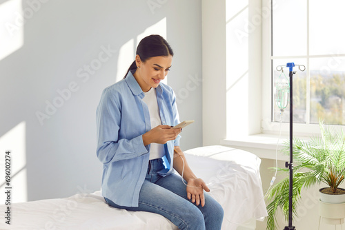 Young happy smiling woman sitting on the couch in medical clinic and using the smartphone while receiving IV drip infusion and vitamin therapy. Female person receiving injection therapy.