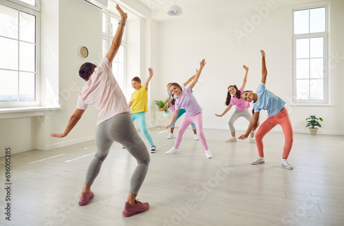Female young choreographer showing her students girls stretching exercises in dance studio. Happy kids doing dance workout. Children training in choreography class, kids sport concept.