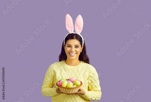 Studio shot of happy Easter bunny girl. Cheerful young woman in yellow jumper and cute rabbit ears standing isolated on purple background, looking at camera, holding bowl of Easter eggs and smiling