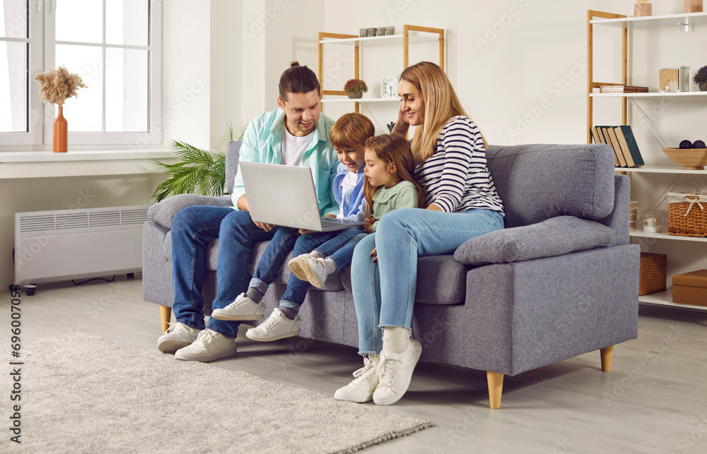 Happy young family with two kids girl and boy sitting on sofa using modern laptop together. Smiling parents resting on couch enjoying weekend watching funny cartoon online or talking on video call.
