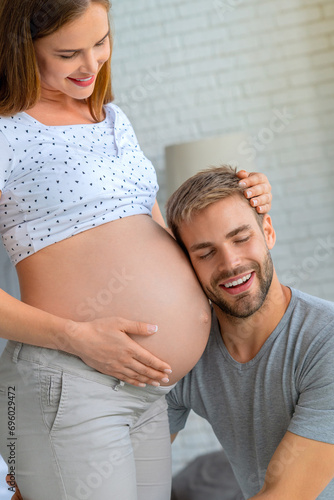Vertical shot of handsome man that listening to his beautiful pregnant wife's tummy and smiling with eyes closed