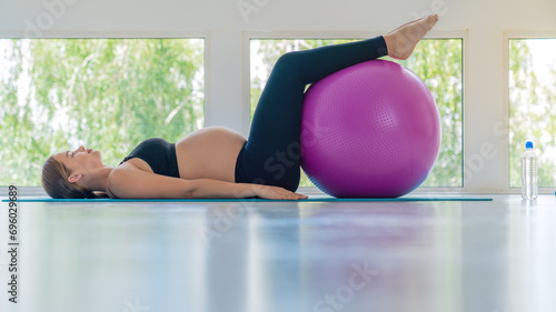 Beautiful active pregnant woman exercising using rubber ball at home against white light window space