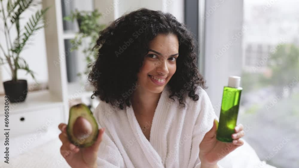 Smiling african woman in after-shower outfit presenting green avocado in fruit and cosmetic oil while sitting behind bed tray at home. Cheerful lady recommending natural ingredients in facial therapy. Smiling african woman in after-shower outfit presenting green avocado in fruit and cosmetic oil while sitting behind bed tray at home. Cheerful lady recommending natural ingredients in facial therapy.