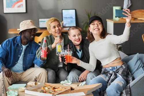 Medium long shot of happy diverse young friends at home taking selfie holding fizzy drinks after lunch with pizza