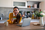 Joyful businesswoman feeling excited holding credit card sitting at desk in the kitchen with laptop screaming with happiness.