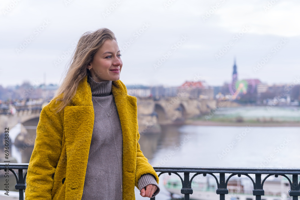 happy beautiful young woman, tourist in a yellow coat looking to the side, standing on a bridge overlooking the city of Dresden, Germany. Travel. New places