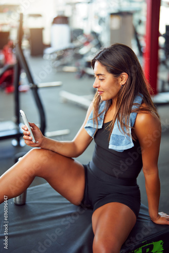 Latin woman sitting in the gym resting between repetitions checking her cell phone 