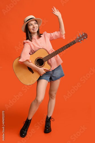 Pretty young woman with acoustic guitar waving hand on orange background
