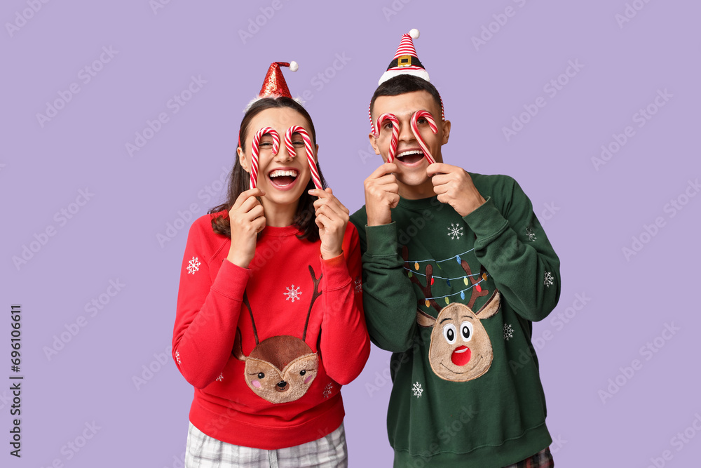 Obraz premium Funny young couple in Christmas pajamas and with candy canes on lilac background