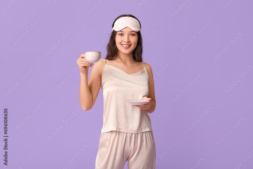 Beautiful young Asian woman in pajamas with sleeping mask and cup of coffee on purple background Beautiful young Asian woman in pajamas with sleeping mask and cup of coffee on purple background