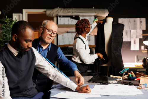 Master tailor working on customer custom made business attire while seamstress does needleworking on sartorial garment. Skilled atelier shop employees helping client during customization comission