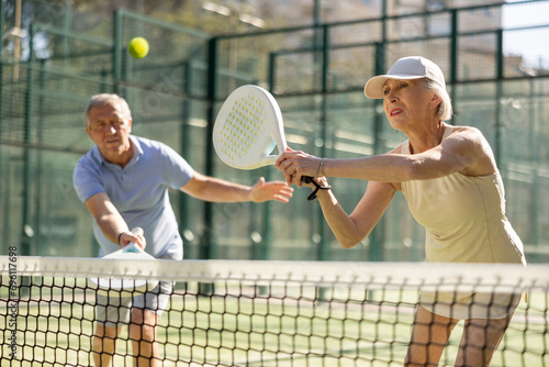 Sportive elderly man and woman engaged in Padel Tennis in open-air court of tennis club © JackF