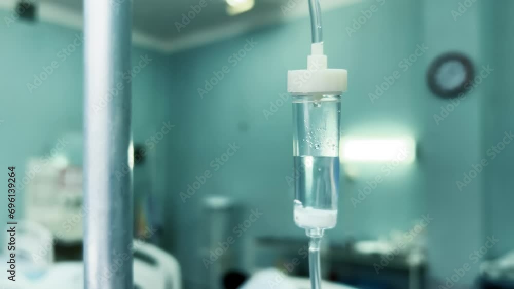 Drip chamber in an IV hanging on a stand against the background of blurred medical beds in a clinic ward. Intravenous injections, medical care in a clinic. Recovering patient in a hospital. Close up Drip chamber in an IV hanging on a stand against the background of blurred medical beds in a clinic ward. Intravenous injections, medical care in a clinic. Recovering patient in a hospital. Close up