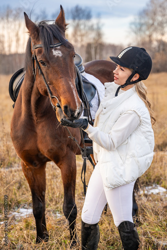 Blond professional female jockey standing near horse in field. Friendship with horse