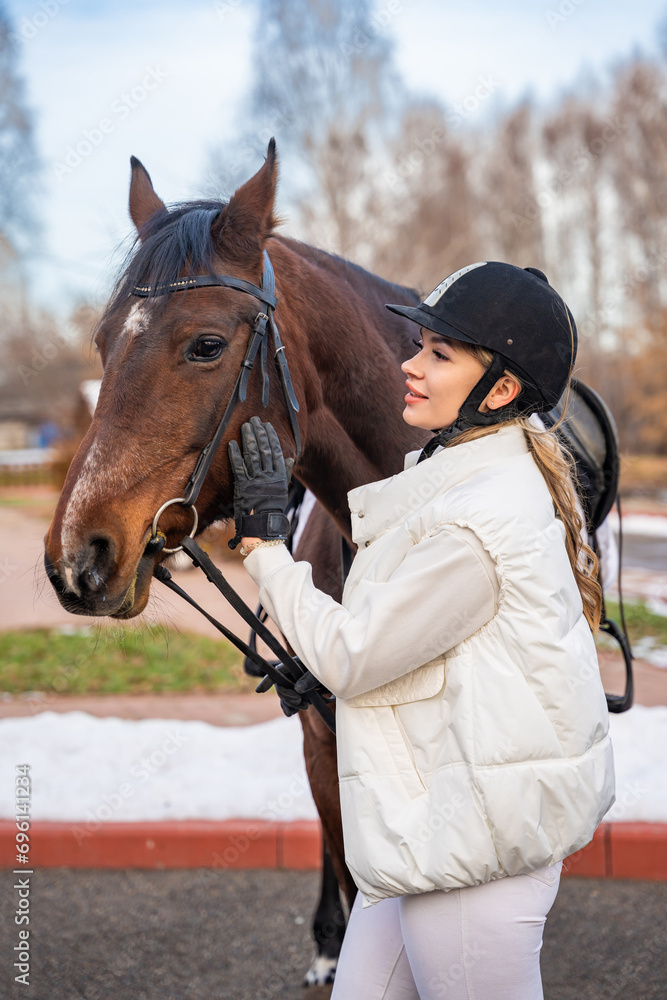 Obraz premium Beautiful blond professional female jockey standing near brown horse on farm. Friendship with horse