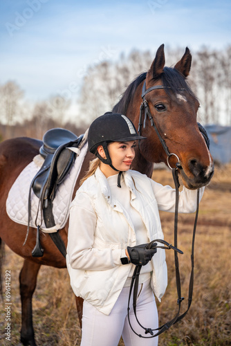 Pretty blond professional female jockey standing near brown horse in field. Friendship with horse