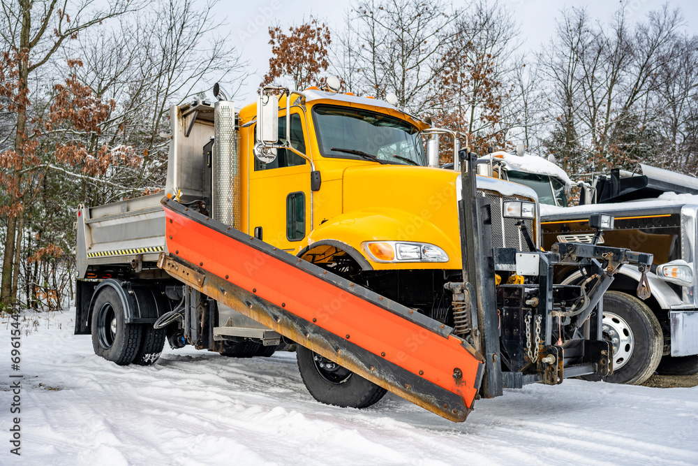 Yellow big rig semi truck tractor with snow shovel and compact dump truck body standing in row on the winter industrial parking lot covered with thick layer of snow Yellow big rig semi truck tractor with snow shovel and compact dump truck body standing in row on the winter industrial parking lot covered with thick layer of snow