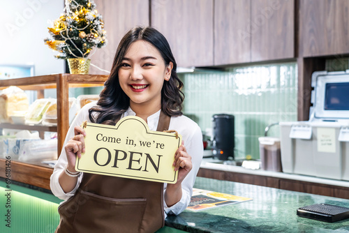 Happy beautiful Asian female waitress or younger shopkeeper in an apron holding open sign while standing at cafe or in restaurant, with pleasured smiling, welcome, business owner startup ideas