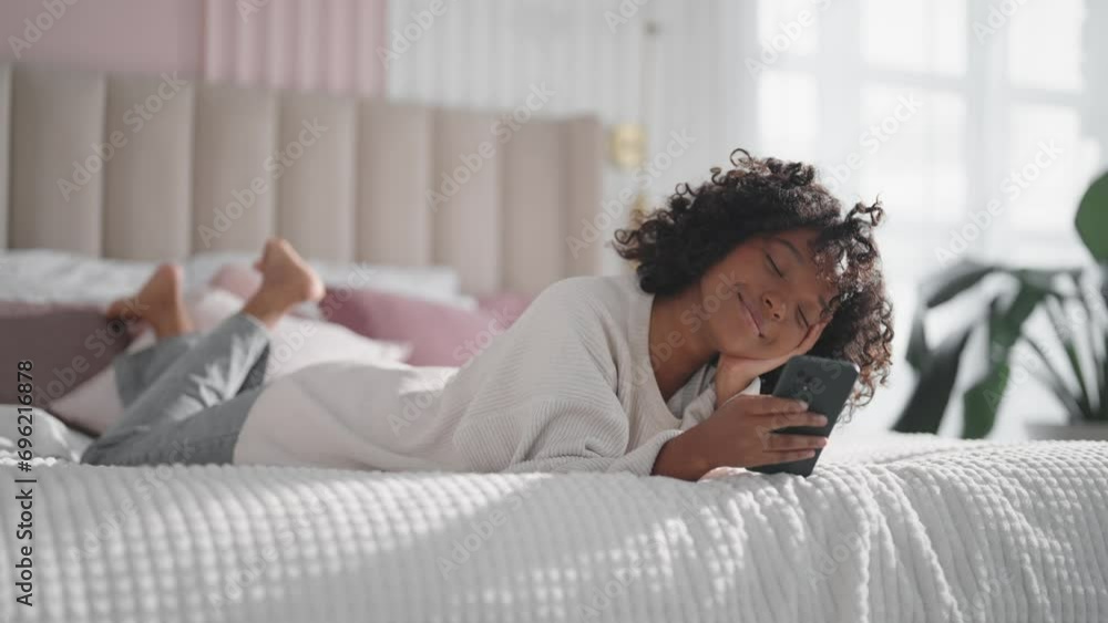 Smiling african american curly woman reading message on smartphone chatting with friend lying in bed at home lazy morning. Positive female enjoying communication in social media looks at phone screen. Smiling african american curly woman reading message on smartphone chatting with friend lying in bed at home lazy morning. Positive female enjoying communication in social media looks at phone screen.