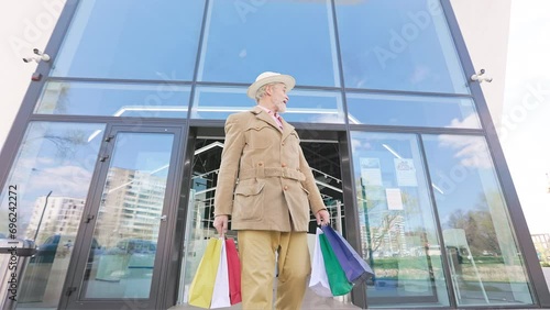 Low angle view contented old man in stylish outfit walking out of mall and carrying multiple shopping bags in hands. Smiling senior male leaving contented with bargain purchases and customer service. photo