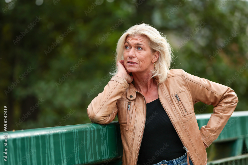 Older blonde woman leans on a bridge railing and looks up Older blonde woman leans on a bridge railing and looks up