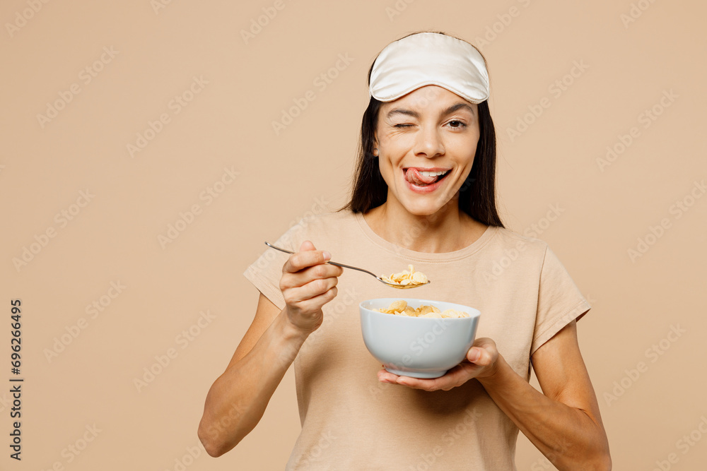 Young Latin woman wears pyjamas jam sleep eye mask rest relax at home eat breakfast muesli cereals with milk fruit in bowl wink isolated on plain beige background studio. Good mood night nap concept. Young Latin woman wears pyjamas jam sleep eye mask rest relax at home eat breakfast muesli cereals with milk fruit in bowl wink isolated on plain beige background studio. Good mood night nap concept.