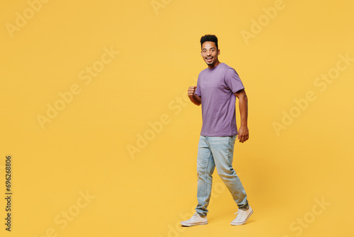 Full body side view smiling young man of African American ethnicity he wears purple t-shirt casual clothes walk go look camera isolated on plain yellow background studio portrait. Lifestyle concept.