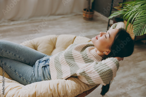 Young woman of Asian ethnicity wear casual clothes holding hands behind neck close eyes sits on armchair stay at home hotel flat rest spend free spare time in living room indoor People lounge concept photo