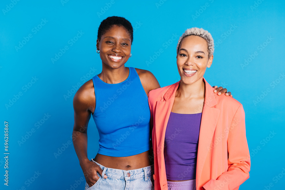 Portrait of a smiling young female couple in front of a blue background at studio. Two women together at studio. Portrait of a smiling young female couple in front of a blue background at studio. Two women together at studio.