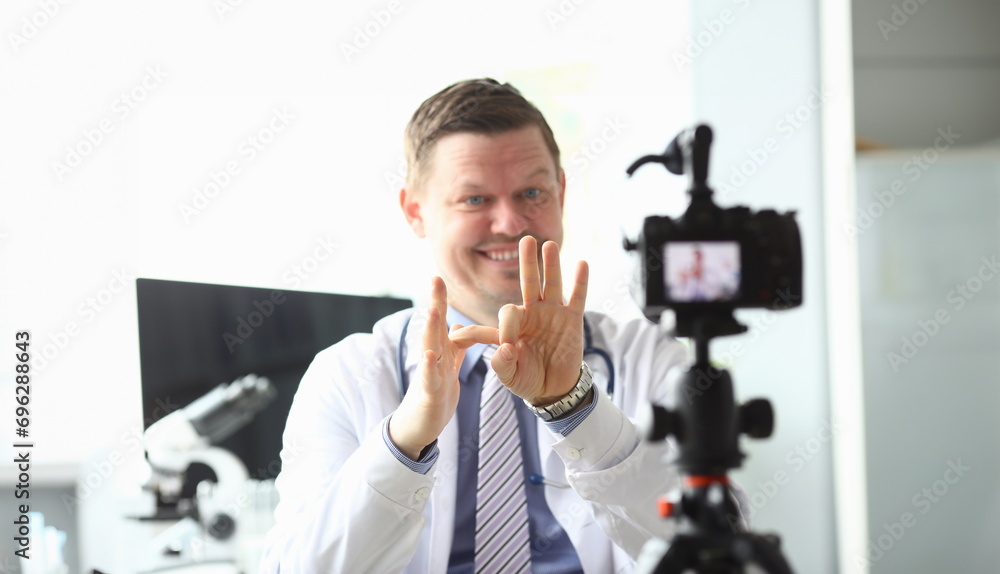 Focus on male hands of handsome doctor showing funny jokes with gestures by hands. Smiling blogger recording popular video content to camera. Blogging concept - obrazy, fototapety, plakaty 