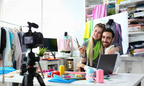Fashion Industry Blog Content Clothes Creation. Blond Seamstress with Scissors and Handsome Caucasian Designer Hugging at Wokplace. Happy Sewing Production Team Recording Vlog at Camera photo