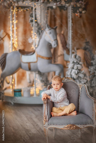 Holiday. New Year. Christmas. Carousel. A boy in a white shirt and beige trousers sits in a chair near gifts and snow-covered Christmas trees. New Year's interior. The kid is standing by the carousel. © Taniya Larina