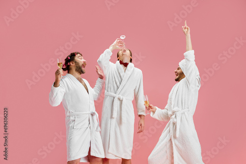 Men, friends in bathrobes and slippers drinking champagne against pink background. Preparation for marriage. Groom's friends. Concept of leisure activity, fun, bachelor party, friendship, spa photo