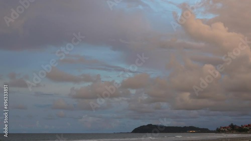 Evening on a tropical beach. There are pinkish cumulus clouds in the light blue sky. The ocean waves roll towards the shore, foaming. A hill and a building with a  light in the distance. Malaysia. 
