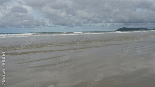 The waves of the turquoise ocean roll towards the shore, foam and bubble on the beach. Clear water spreads in front of the camera. A hill against a blue sky and clouds in the distance. Malaysia.Borneo