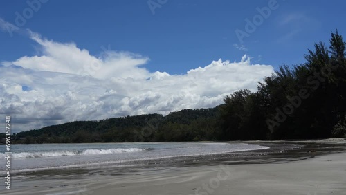 Tropical seascape. The waves of the turquoise ocean roll towards the shore, foaming. The cottages are located in a thicket of green trees on the coast. Clouds in the blue sky. Reflection on wet sand.