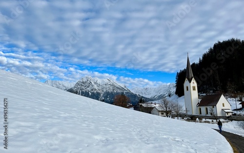 Spaziergang im Winter bei Schnee und blauem Himmel mit Wolken in den Alpen bzw. Bergen bei einer Kirche bei Oberstdorf in Bayern 