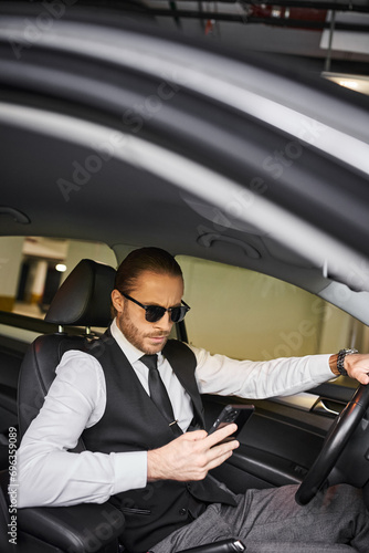 appealing red haired man with beard and sunglasses looking at phone behind steering wheel, business © LIGHTFIELD STUDIOS