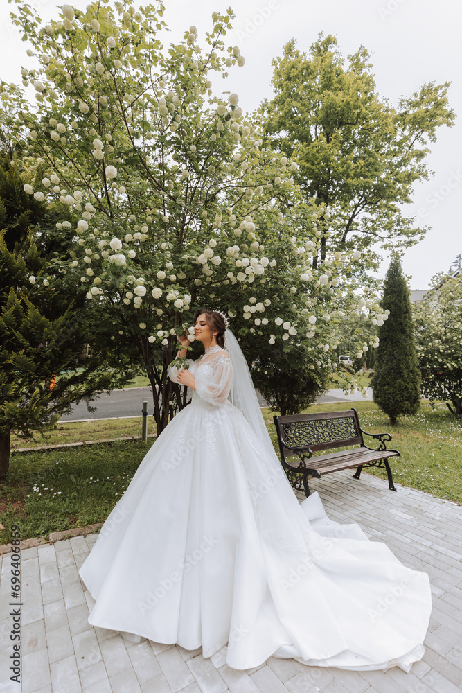 Curly brunette bride in a lace dress with an open bust, in a lush veil holds a bouquet and poses against the background of green trees. Spring wedding