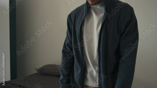 Midsection cropped shot of unrecognizable man putting on dark sweat jacket on white t-shirt and zipping it up while dressing up in bedroom photo